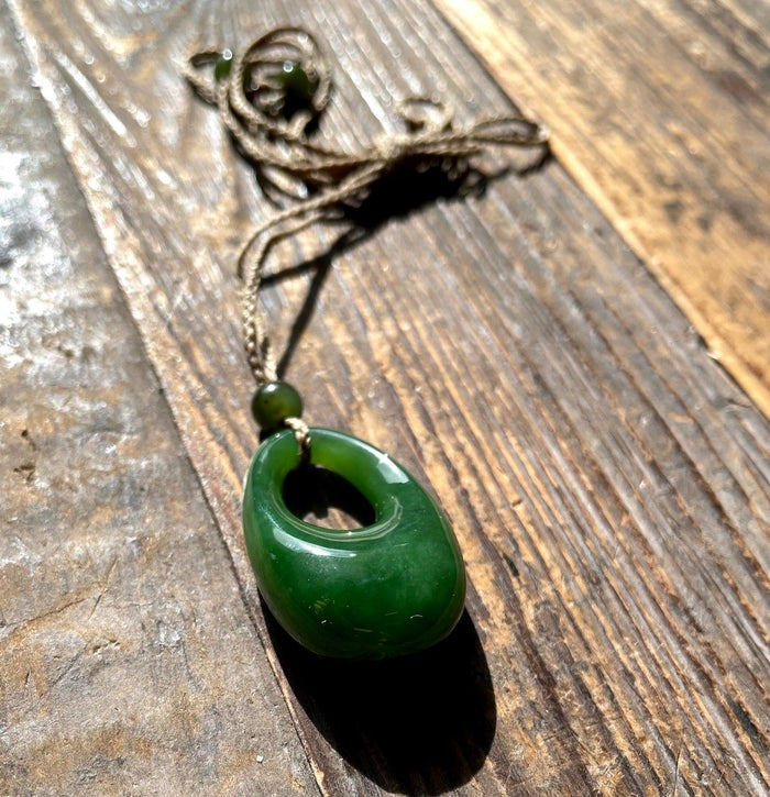 Close up of a jade donut pendant threaded on brown twine with green beads, on a weathered wooden surface.