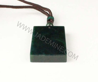 Dark green rectangular jade pendant with brown cord and bead resting on a white surface.