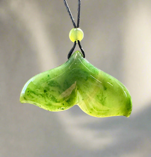 Light green jade whale tail pendant with darker marbling on a black cord, accented by a small yellow bead above.