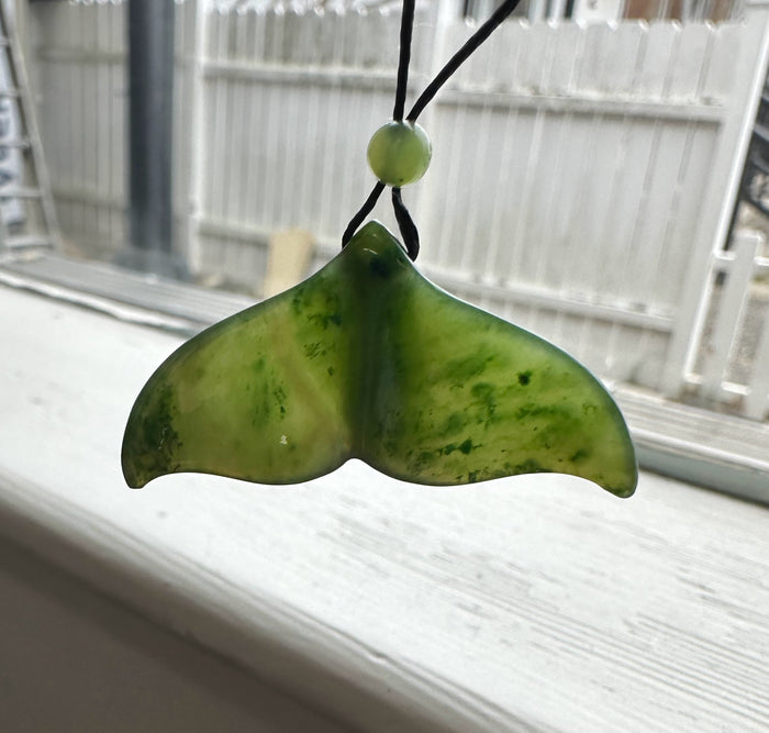 Green jade whale tail pendant on a black cord with a bead, photographed against a bright window.