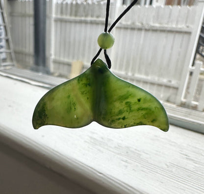 Green jade whale tail pendant on a black cord with a bead, photographed against a bright window.