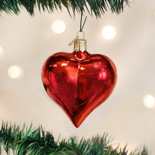 A shiny red heart-shaped ornament hangs from a green Christmas tree branch, with soft white lights blurred in the background.