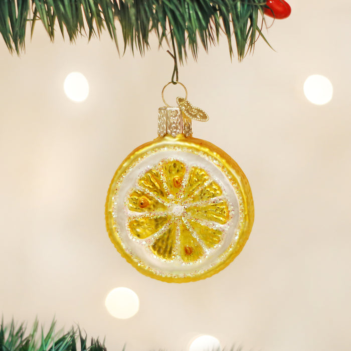 A Christmas tree ornament shaped like a lemon slice hangs from a green branch, with a blurred background of white lights.