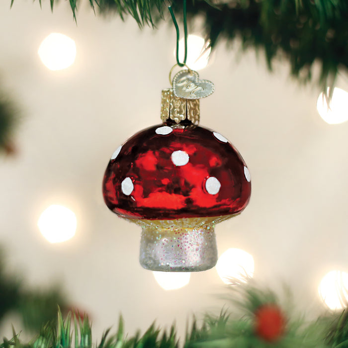 A red and white mushroom-shaped ornament hangs from a Christmas tree branch, surrounded by green needles and soft, out-of-focus holiday lights in the background.