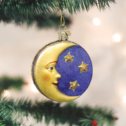 A Christmas tree ornament shaped like a crescent moon with a face, partly covered by a blue section with three gold stars, hangs from a green tree branch with blurred white lights in the background.