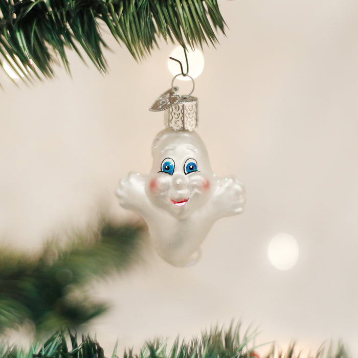 A white glass ornament shaped like a smiling ghost with blue eyes and rosy cheeks hangs from a green Christmas tree branch against a softly blurred background.