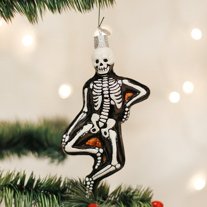 A glass Christmas tree ornament shaped like a skeleton, with one arm on its hip and one leg crossed, hangs from a green tree branch. The background features soft, out-of-focus white lights.