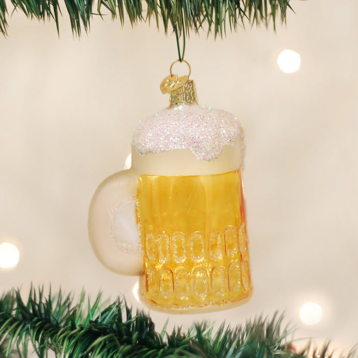 A Christmas tree ornament shaped like a frothy beer mug, decorated with glitter to look like foam, hangs from a green Christmas tree branch with blurred lights in the background.