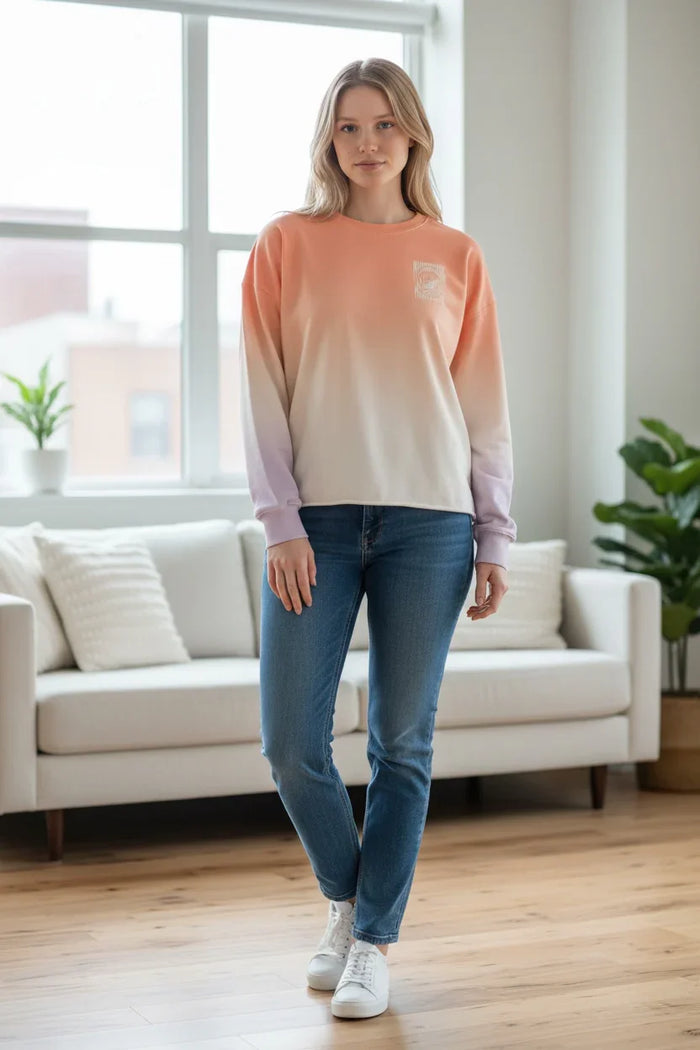 Ombre peach to lavender surf sweatshirt with subtle logo, paired with blue jeans and white sneakers.