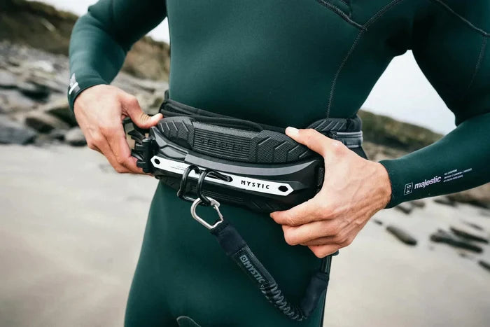 Mystic kitesurf harness with black and silver design, worn with green wetsuit on beach