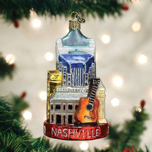 A colorful Christmas tree ornament featuring Nashville landmarks, a guitar, and the words â€œGrand Ole Opryâ€_x009d_ and â€œNashville,â€_x009d_ hanging on a green tree with blurred lights in the background.