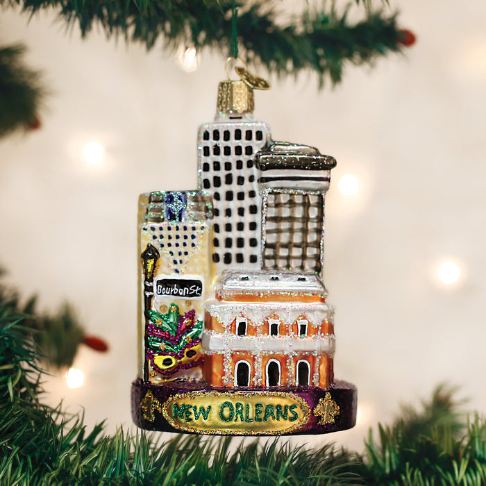 A Christmas tree ornament featuring miniature New Orleans landmarks, including buildings, a Bourbon Street sign, and a Mardi Gras mask, with New Orleans written at the base, hanging on a tree branch.