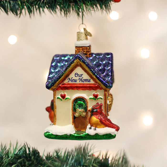 A glass Christmas ornament shaped like a house with â€œOur New Homeâ€_x009d_ written on the roof, decorated with two red cardinals and snow, hangs on a Christmas tree with blurred lights in the background.
