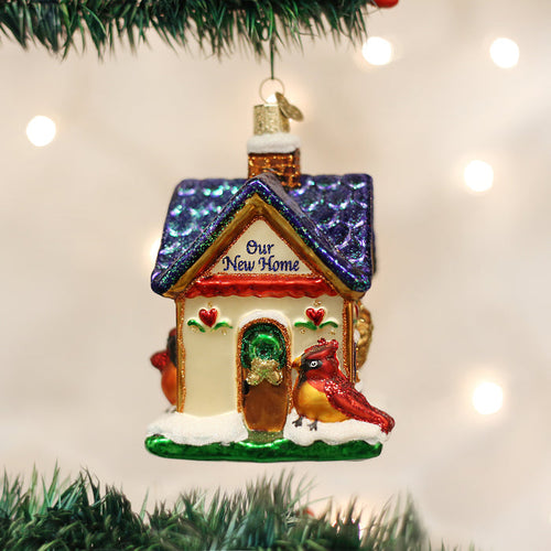 A glass Christmas ornament shaped like a house with â€œOur New Homeâ€_x009d_ written on the roof, decorated with two red cardinals and snow, hangs on a Christmas tree with blurred lights in the background.