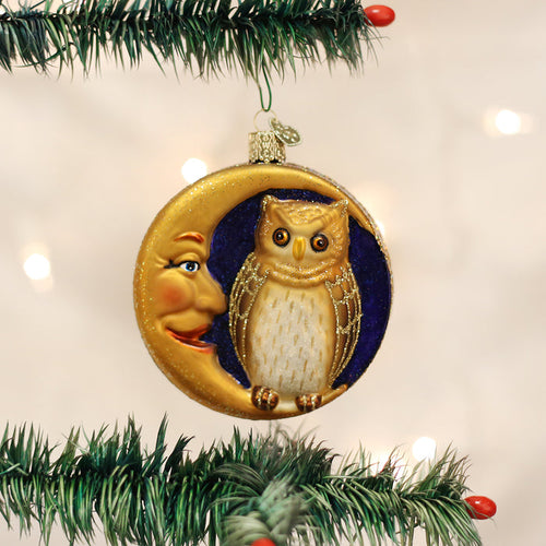 A Christmas tree ornament featuring a crescent moon with a smiling face, embracing a detailed owl against a dark blue background, hangs on a pine branch with blurred lights in the background.