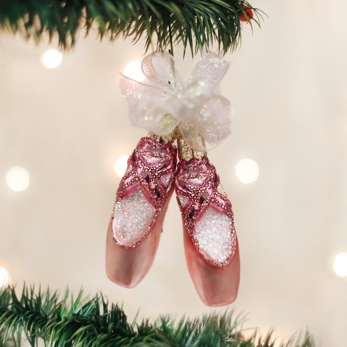 A pair of pink glittery ballet slipper ornaments with bows and a sheer ribbon, hanging from a Christmas tree branch with blurred lights in the background.