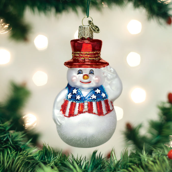 A snowman ornament with a red, white, and blue patriotic vest and top hat hangs on a Christmas tree, surrounded by blurred lights in the background.