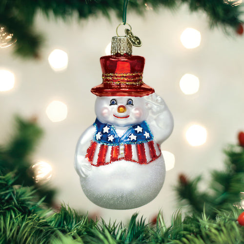 A snowman ornament with a red, white, and blue patriotic vest and top hat hangs on a Christmas tree, surrounded by blurred lights in the background.