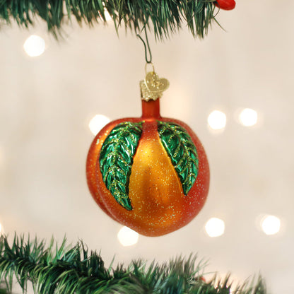 A peach-shaped Christmas ornament with green leaves and gold glitter hangs from a green Christmas tree branch with blurred white lights in the background.