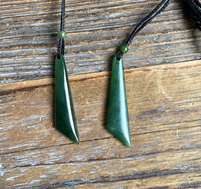 Two green jade drop pendants on black cords with small green beads against a wooden background