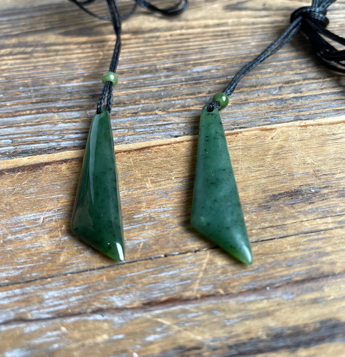Two green jade drop pendants on black cords with beads placed on a wooden surface