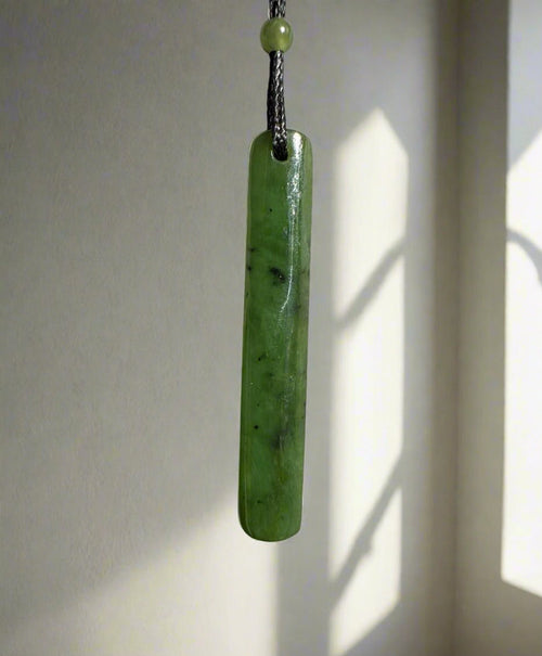 Close-up of a green jade drop pendant on a black cord hanging against a light colored wall.
