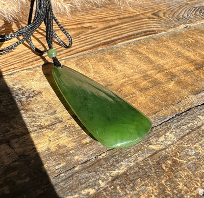 Green jade teardrop pendant on black cord with a small bead above, laid on a wood surface