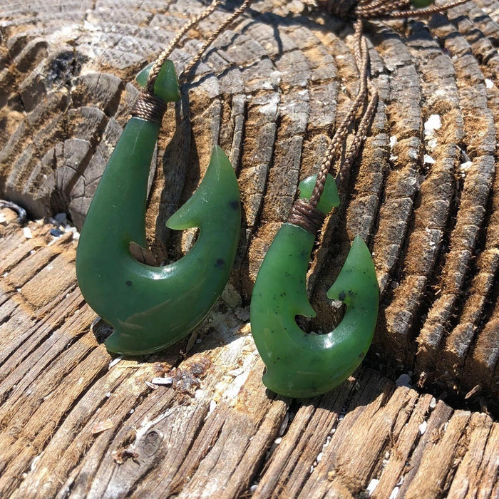 Two green jade fish hook pendants with braided cords resting on weathered wood