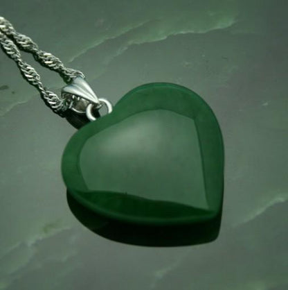 Close-up of a polished green jade heart pendant with a silver bail resting on a dark green surface.