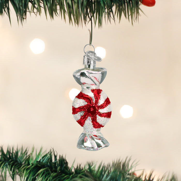 A Christmas tree ornament shaped like a red and white peppermint candy hangs from a green tree branch with blurred white lights in the background.