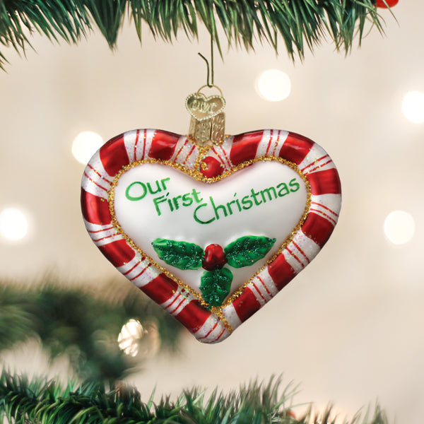 A heart-shaped Christmas ornament with red and white stripes hangs on a tree. It reads â€œOur First Christmasâ€_x009d_ in green and features holly with red berries at the bottom.