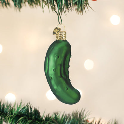 A green glass ornament shaped like a pickle hangs from a decorated Christmas tree, with soft, out-of-focus lights in the background.