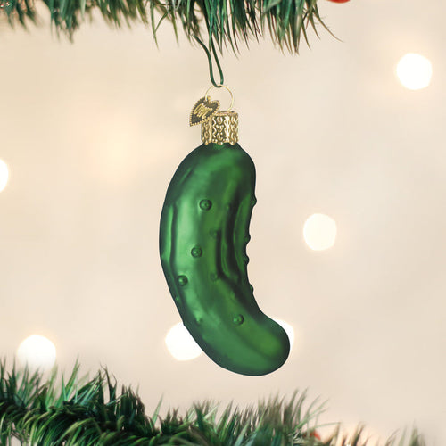 A green glass ornament shaped like a pickle hangs from a decorated Christmas tree, with soft, out-of-focus lights in the background.