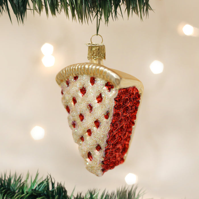 A Christmas tree ornament shaped like a slice of cherry pie, decorated with red and white glitter, hangs from a green tree branch with blurred lights in the background.