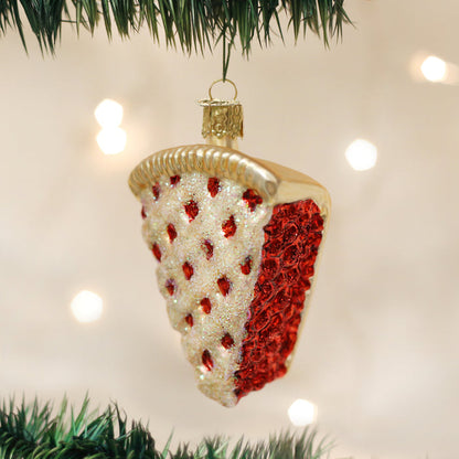 A Christmas tree ornament shaped like a slice of cherry pie, decorated with red and white glitter, hangs from a green tree branch with blurred lights in the background.