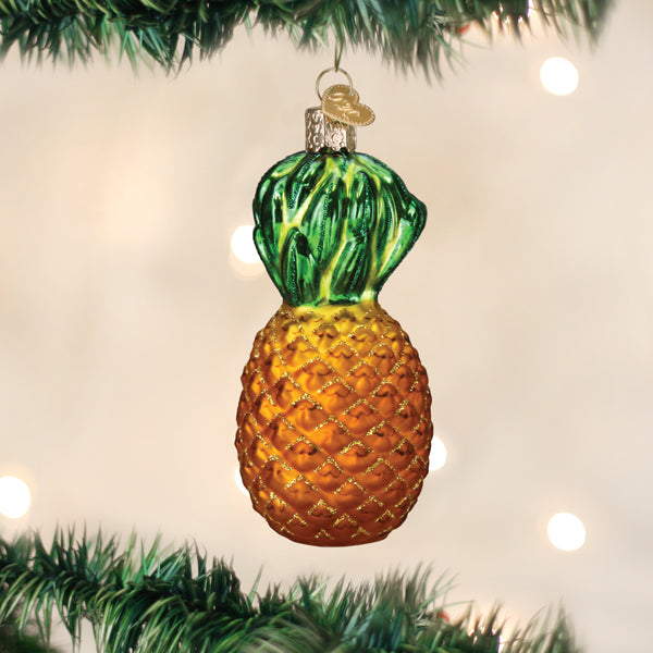 A shiny glass Christmas ornament shaped like a pineapple hangs from a green tree branch with blurred white lights in the background.