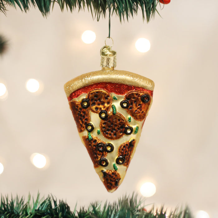 A Christmas tree ornament shaped like a slice of pepperoni pizza with black olives and green peppers, hanging from a tree branch with blurred lights in the background.