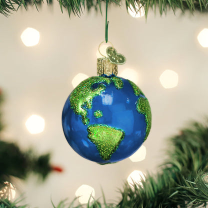 A Christmas tree ornament shaped like a globe, with blue oceans and green glitter continents, hangs among pine branches with soft, glowing lights in the background.