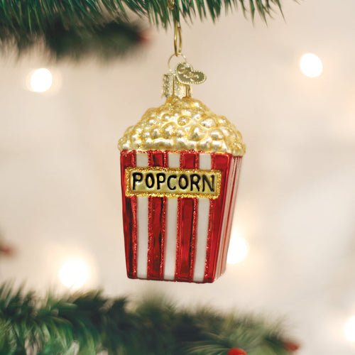 A shiny Christmas tree ornament shaped like a red and white striped popcorn box with gold popcorn on top, hanging on a green branch with soft white lights in the background.