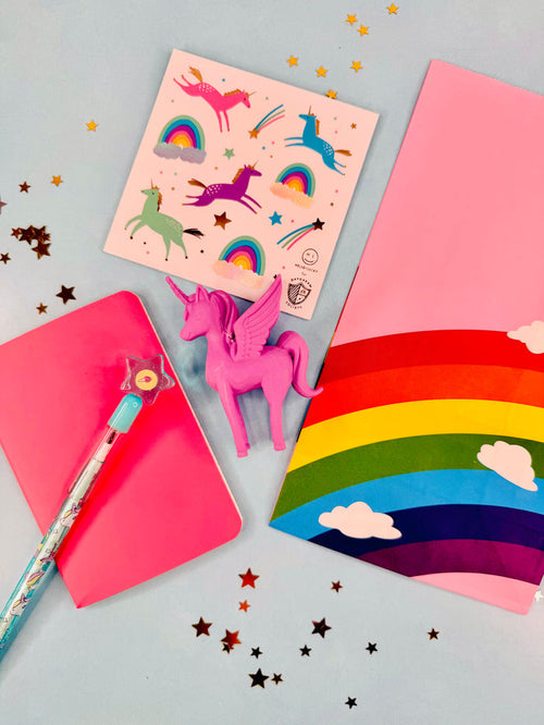 Colorful stationery items including a pink notebook, rainbow card, and unicorn-themed stationery on a gray background.