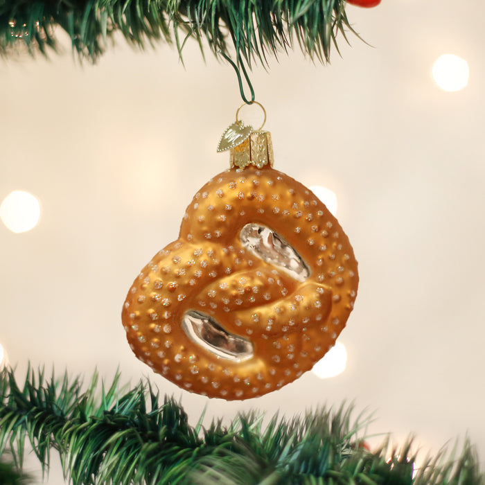 A Christmas ornament shaped like a pretzel hangs from a green tree branch, with a soft, blurred background of white and yellow lights.