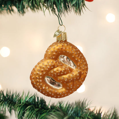 A Christmas ornament shaped like a pretzel hangs from a green tree branch, with a soft, blurred background of white and yellow lights.