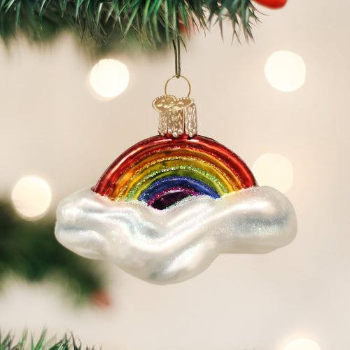 A Christmas tree ornament shaped like a rainbow emerging from white, glittery clouds hangs from a green branch with blurred white lights in the background.