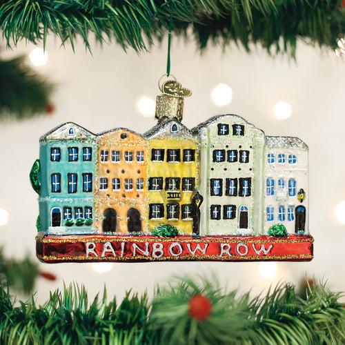 A colorful Christmas tree ornament shaped like a row of pastel houses labeled â€œRainbow Row,â€_x009d_ hanging among green branches with blurred lights in the background.
