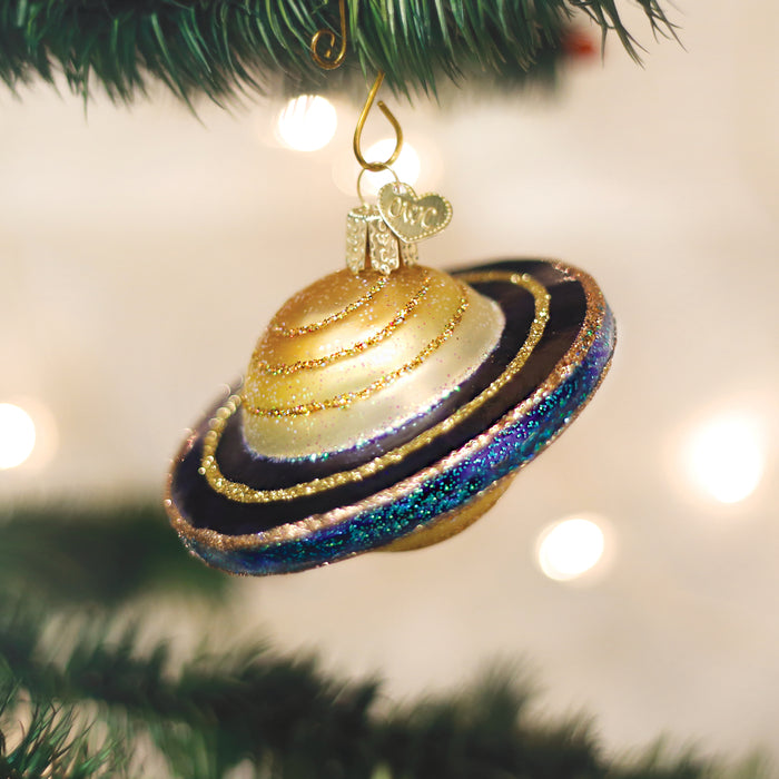 A Christmas tree ornament shaped like the planet Saturn, decorated with gold glitter and colorful rings, hangs from a green pine branch with blurred white lights in the background.
