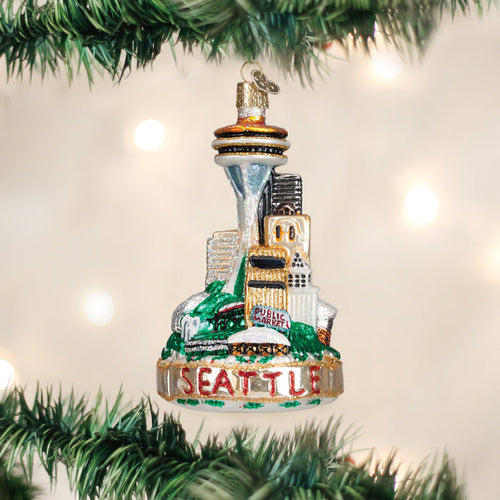 A Christmas tree ornament featuring Seattle landmarks, including the Space Needle and Pike Place Market, hangs on a green tree branch with blurred white lights in the background. Seattle is written at the base of the ornament.