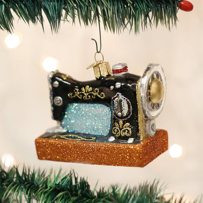 A Christmas tree ornament shaped like a vintage black and gold sewing machine with glittery blue and orange accents hangs among green pine branches.
