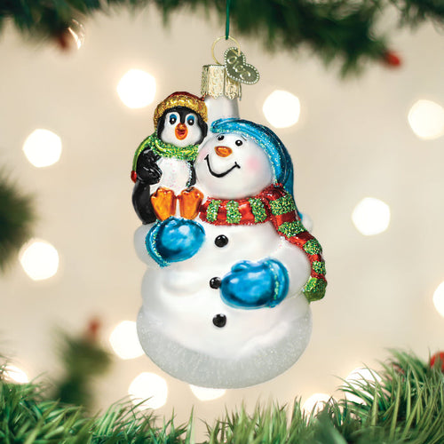 A festive ornament of a smiling snowman in blue mittens and scarf, holding a cheerful penguin, hangs on a Christmas tree with green branches and blurred white lights in the background.