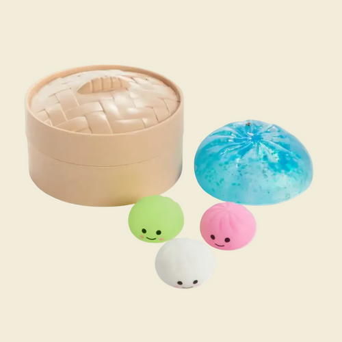 A set of cute bun-shaped squishy toys and a matching storage container. The set includes a beige woven-texture round container, a large blue glittery bun, a green smiley-face bun, a pink pleated bun, and a white bun, all on a solid white background.