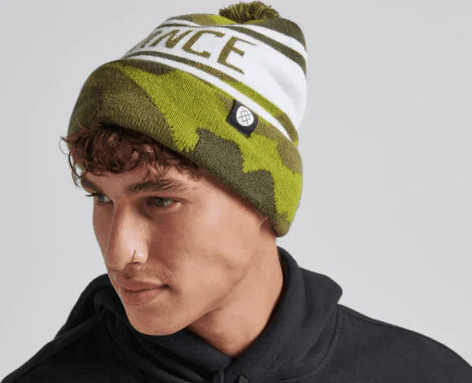 Green camo beanie with white stripe and pom-pom, featuring brand logo, ideal for watersports
